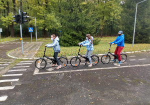 Troje uczniów siedzących na rowerach oczekuje na sygnał egzaminatora do rozpoczęcia jazdy.